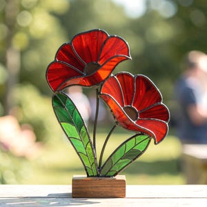 Könnte beinhalten: Eine Buntglasskulptur mit zwei leuchtend roten Mohnblumen mit dunklen Zentren, die von grünen Glasblättern und Stielen getragen werden. Das florale Stück ist auf einem rechteckigen Holzsockel montiert und zeigt eine kunstvolle Bleiverglasung.