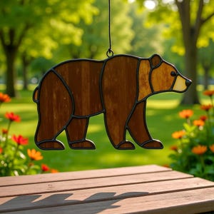 May include: A stained glass bear suncatcher with a brown body and a lighter brown head and snout. The bear is outlined in black and hangs from a black string. The background is a blurred view of a garden with trees and flowers.