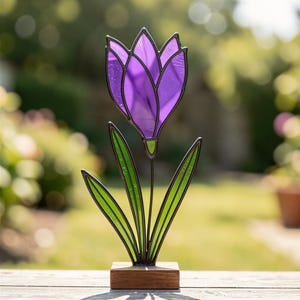 Puede incluir: Una escultura de tulipán en vitral que presenta una cabeza de flor de color púrpura vibrante con líneas de plomo oscuras. La flor tiene tres hojas de vidrio verde largas y puntiagudas que salen de un tallo central, sobre una base de madera.