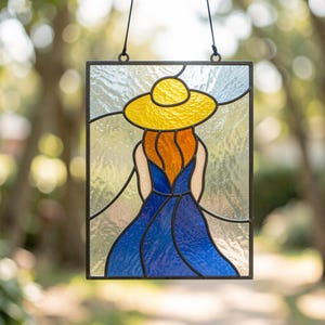 May include: A rectangular stained glass suncatcher featuring a figure viewed from behind. The figure wears a bright yellow sun hat, has orange hair, and is dressed in a flowing blue dress. The background consists of clear, textured glass panels.