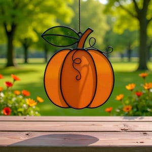 May include: An orange stained glass pumpkin decoration with a black metal outline, stem, and leaf. The pumpkin is hanging in front of a blurred green background with flowers and trees. A decorative item for autumn.