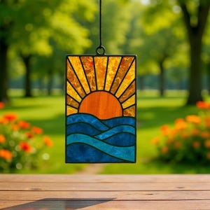 Puede incluir: Un panel rectangular de vidrieras que representa una puesta de sol sobre el océano. El sol es naranja, con rayos amarillos y naranjas. El océano está representado por olas azules y turquesas. El panel está enmarcado en negro y cuelga de una cadena negra.