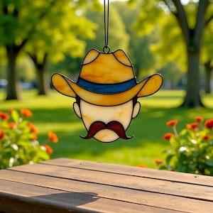 May include: Stained glass cowboy face ornament with a yellow hat, blue hatband, and a red mustache. The ornament is hanging in front of a blurred green background with a wooden table in the foreground.