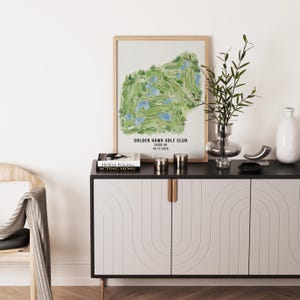 May include: A framed watercolor print of the Golden Hawk Golf Club in Casco, MI, featuring a green and blue course layout. The print is displayed on a black and white cabinet with gold handles, alongside decorative items and a book.