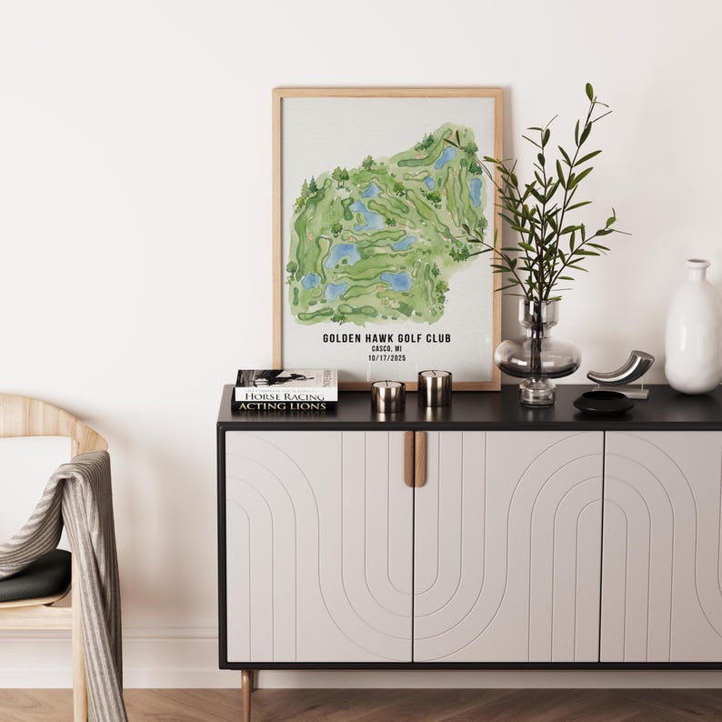 May include: A framed watercolor print of the Golden Hawk Golf Club in Casco, MI, featuring a green and blue course layout. The print is displayed on a black and white cabinet with gold handles, alongside decorative items and a book.
