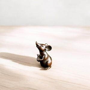 Peut inclure: Une figurine de souris de couleur bronze, la tête levée et les yeux fermés. La souris tient un petit objet dans ses pattes. La figurine est posée sur une surface en bois de couleur claire.
