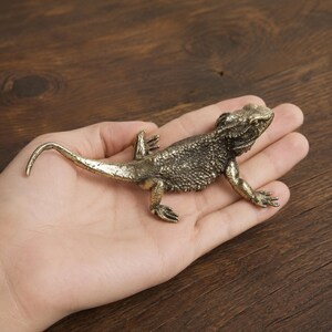 May include: A small, detailed bronze-colored metal figurine of a bearded dragon lizard. The sculpture features a textured, scaly surface, a long curved tail, and four legs positioned as if the lizard is crawling across a flat surface.
