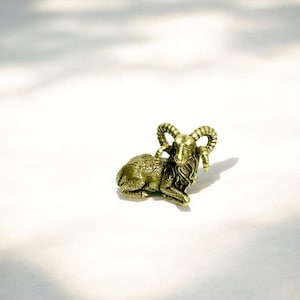 Könnte beinhalten: Kleine, detaillierte Widderfigur aus Messing in liegender Pose. Der Widder hat große, gebogene Hörner und ein strukturiertes Fell. Die Figur steht vor einem schlichten, weißen Hintergrund mit Schatten.