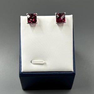 May include: A pair of square-cut, deep red gemstone stud earrings. The earrings are set in a silver-colored metal and displayed on a white and blue jewelry display stand. The gemstones have a faceted appearance, catching the light.