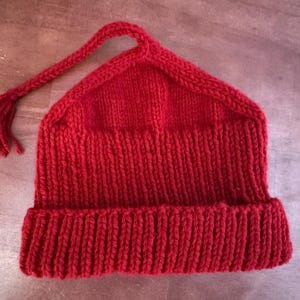 May include: A vibrant red knit hat with a unique pointed top and a long, braided tassel. The hat features a folded brim and is made of a thick, textured yarn. This cozy hat is perfect for cold weather.