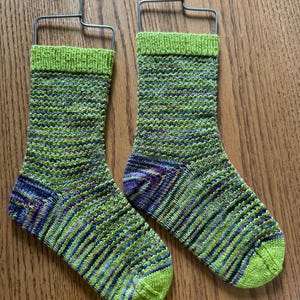May include: A pair of hand-knitted socks in shades of green, blue, and purple. The socks have a ribbed cuff and toe, and are displayed on metal hangers. The yarn has a variegated pattern.