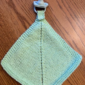 May include: A light green square-shaped knitted security blanket featuring a textured border and a central vertical seam. A pale blue pacifier is attached to the top of the blanket by a small knitted loop, creating a unified baby accessory.