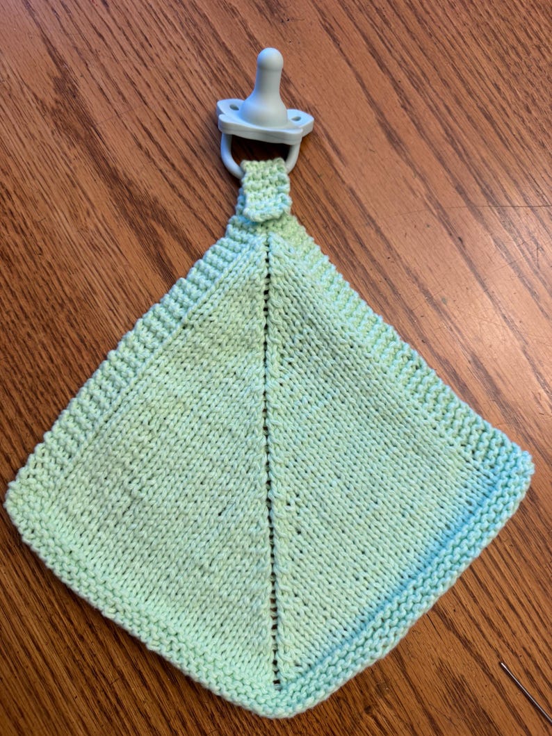 May include: A light green square-shaped knitted security blanket featuring a textured border and a central vertical seam. A pale blue pacifier is attached to the top of the blanket by a small knitted loop, creating a unified baby accessory.