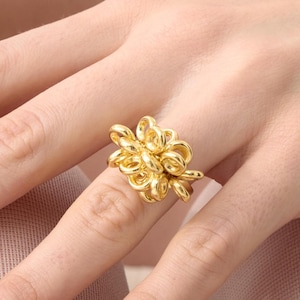 May include: A gold ring with a floral design. The ring is worn on a finger, and the design features multiple flower-like elements clustered together. The ring's intricate details are visible, and the overall aesthetic is elegant and decorative.