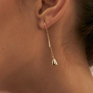 May include: A close-up of a person wearing a gold earring. The earring features a long, slender chain with a small, bell-shaped charm. The person is wearing a silver pleated top.