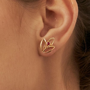 May include: A gold-colored stud earring with a floral design. The earring features three stylized petal shapes surrounding a small, round, red gemstone. The earring is worn on an ear.