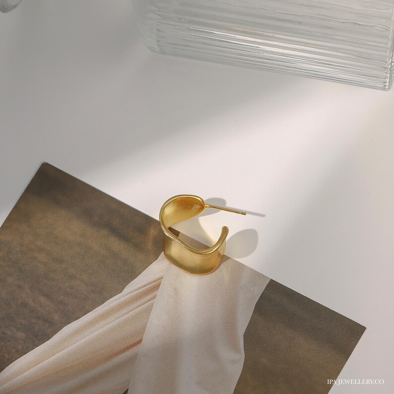 May include: A gold-toned, open hoop earring with a slightly curved, flat design. The earring is made of metal and has a smooth, polished finish.
