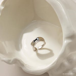 May include: A silver ring with a unique, curved design. The ring is shown in a white ceramic bowl.
