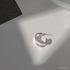 May include: A silver hoop earring with a unique, curved, and slightly asymmetrical design. The earring is made of a smooth, polished metal.