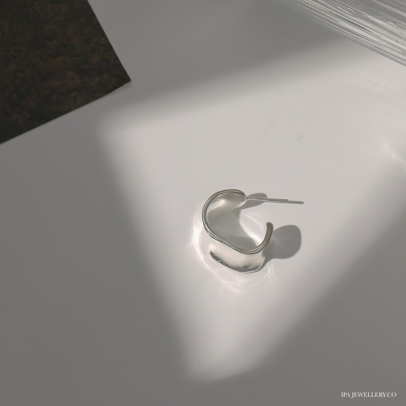 May include: A silver hoop earring with a unique, curved, and slightly asymmetrical design. The earring is made of a smooth, polished metal.