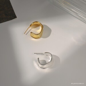 May include: Two pairs of hoop earrings, one gold and one silver. The earrings are made of a smooth, curved metal and have a simple, modern design.