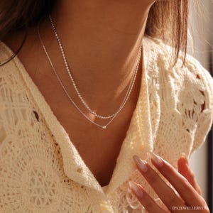 May include: A silver chain necklace with a small silver bead pendant. The necklace is layered with a second, thinner silver chain.