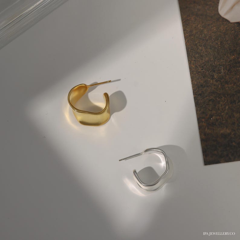 May include: Two pairs of earrings, one gold and one silver, with a unique, asymmetrical, wavy design. The earrings are simple and modern.