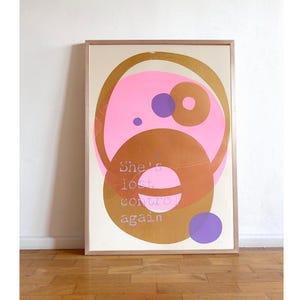 Könnte beinhalten: Gerahmter Kunstdruck mit einem abstrakten Design. Das Kunstwerk zeigt überlappende Formen in Rosa, Gold und Lila auf cremefarbenem Hintergrund. Der Text lautet "She's lost control again". Der Druck ist in einem Holzrahmen.