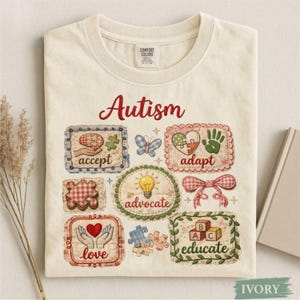 以下が含まれることがあります： アイボリー色のTシャツ。赤い筆記体で「Autism」と書かれています。シャツには、「accept」、「adapt」、「love」などの言葉が書かれた様々なパッチ、パズルのピース、蝶、電球が描かれています。