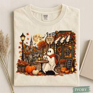Może przedstawiać: T-shirt w kolorze kości słoniowej z grafiką przedstawiającą ducha siedzącego przy kawiarnianym stoliku i pijącego kawę. Scena zawiera pełnię księżyca, jesienne liście, dynie, rower oraz szyld z napisem Moonlit Beans Cafe i Coffee Books Autumn.