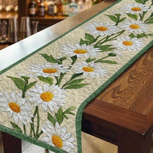 May include: A quilted table runner featuring a daisy floral design. The runner has a cream-colored background with white daisies, yellow centers, and green leaves. A green border frames the edges. The runner is displayed on a wooden table.