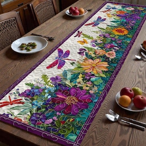 Puede incluir: Un camino de mesa floral con flores coloridas, libélulas y mariposas sobre un fondo blanco. El camino de mesa, con borde morado, está colocado sobre una mesa de madera. El diseño incluye tonos de morado, naranja y verde.
