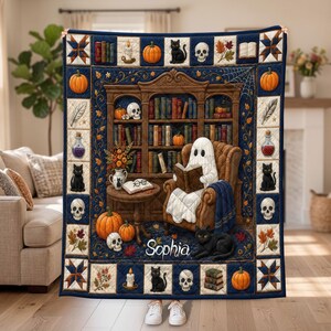 May include: A rectangular quilt featuring a central scene of a ghost reading a book in an armchair within a library, surrounded by pumpkins, skulls, and black cats. The border includes patterned squares and the name Sophia in white script text.