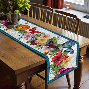 May include: A rectangular table runner with a white background and a floral and butterfly design. The runner features a teal border and is decorated with colorful flowers and butterflies in shades of red, orange, pink, yellow, and blue.