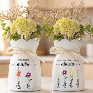 Könnte beinhalten: Zwei weiße Keramikvasen mit Blumenarrangements. Jede Vase hat einen gewellten Rand, ein gebundenes Band und Text. Eine Vase trägt die Aufschrift "madre" mit den Namen "Gera" und "Lili". Die andere trägt die Aufschrift "abuela" mit den Namen "Cristofer", "Kevin", "Johnny" und "Alonso".