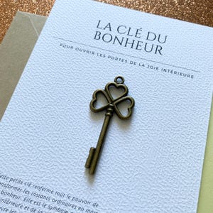 Peut inclure: Une breloque clé en métal couleur bronze avec un anneau en forme de trèfle composé de trois cœurs. La clé est fixée sur une carte blanche texturée portant le texte LA CLÉ DU BONHEUR et POUR OUVRIR LES PORTES DE LA JOIE INTÉRIEURE.