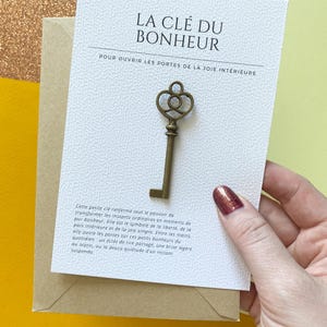 Peut inclure: Une clé en métal de couleur bronze avec un anneau décoratif fixée sur une carte blanche texturée. La carte porte le texte LA CLÉ DU BONHEUR et un paragraphe en français. La clé est centrée sur la carte posée sur une enveloppe brune.