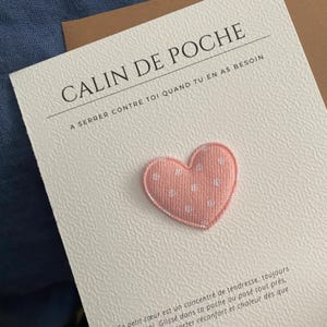 Peut inclure: Une carte blanche texturée portant le texte CALIN DE POCHE et A SERRER CONTRE TOI QUAND TU EN AS BESOIN. Un petit cœur en tissu rose rembourré avec un motif à pois blancs est fixé au centre de la carte au-dessus d'un texte français additionnel.