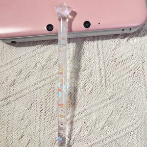 Peut inclure: Un stylet transparent avec une étoile scintillante au sommet et des paillettes colorées à l'intérieur. Le stylet repose sur une console de jeu portable rose, sur un fond en tricot blanc.