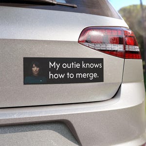 May include: A black bumper sticker with a woman's face and the text "My outie knows how to merge."