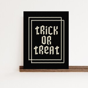 May include: A black framed art print with the words "TRICK OR TREAT" in white, stylized text. The text is centered within a white beaded border on a black background. The print is displayed on a wooden shelf.