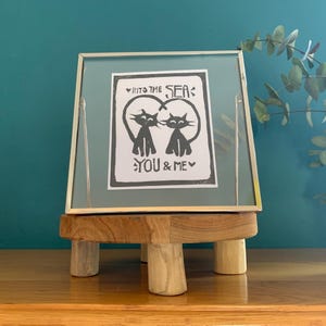 Può includere: Una stampa incorniciata con un'illustrazione in bianco e nero di due gatti che formano un cuore con le loro code. Il testo dice "Into the Sea: You & Me".
