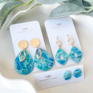 May include: A collection of handmade polymer clay earrings in shades of blue and turquoise. The earrings include teardrop, diamond, and round stud styles, with gold-colored accents. The brand name "Everbryn Clay" is visible on the product cards.