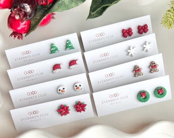 May include: A collection of Christmas-themed stud earrings on white cards. Designs include Christmas trees, Santa hats, snowmen, bows, snowflakes, gingerbread men, poinsettias, and wreaths. The cards display the text "EVERBRYN CLAY designs".