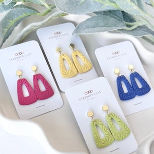 May include: A collection of handcrafted clay earrings in various colors, including pink, yellow, blue, and green. Each pair features a unique geometric design with gold-colored posts and is displayed on a white card. The brand name "EVERBRYN CLAY" is printed on the cards.