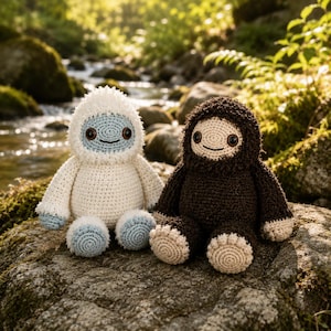 May include: Two crocheted plush creatures sitting side by side. The left figure is white with a light blue face and feet. The right figure is dark brown with beige feet. Both have round dark eyes, simple stitched smiles, and textured yarn bodies.