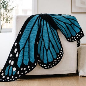 K&ouml;nnte beinhalten: Eine geh&auml;kelte Decke in Form eines gro&szlig;en Schmetterlingsfl&uuml;gels mit einem leuchtend t&uuml;rkisfarbenen und schwarzen Farbschema sowie wei&szlig;en Punkten an den R&auml;ndern. Das Textildesign ahmt die komplexen Adernmuster eines Schmetterlingsfl&uuml;gels nach.