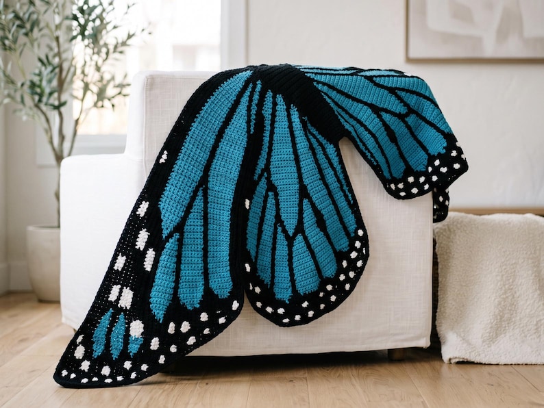 K&ouml;nnte beinhalten: Eine geh&auml;kelte Decke in Form eines gro&szlig;en Schmetterlingsfl&uuml;gels mit einem leuchtend t&uuml;rkisfarbenen und schwarzen Farbschema sowie wei&szlig;en Punkten an den R&auml;ndern. Das Textildesign ahmt die komplexen Adernmuster eines Schmetterlingsfl&uuml;gels nach.
