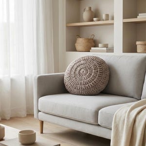 Puede incluir: Una escena de sal&oacute;n de tonos neutros con un sof&aacute; gris claro y un coj&iacute;n redondo de ganchillo. Una manta color crema est&aacute; sobre el sof&aacute;. Estanter&iacute;as en el fondo exhiben objetos decorativos, incluyendo jarrones y cestas. Cortinas blancas transparentes enmarcan la escena.