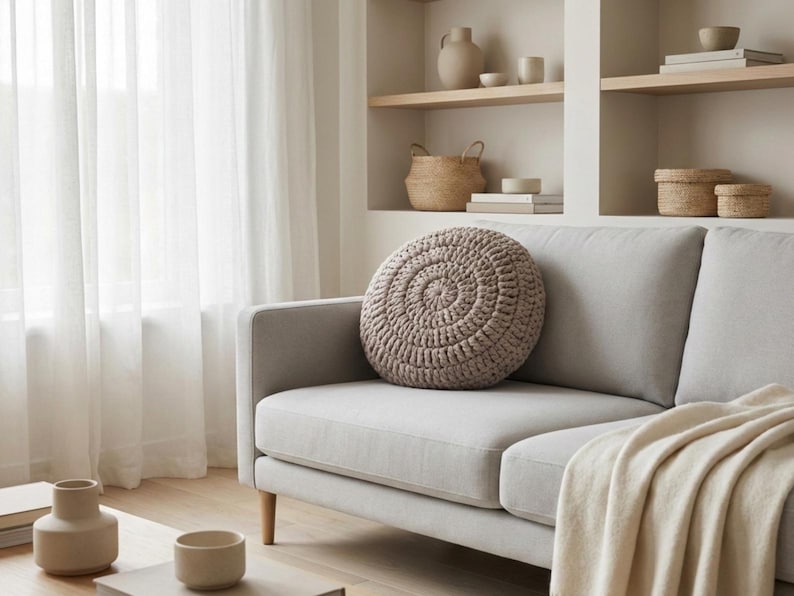 Puede incluir: Una escena de sal&oacute;n de tonos neutros con un sof&aacute; gris claro y un coj&iacute;n redondo de ganchillo. Una manta color crema est&aacute; sobre el sof&aacute;. Estanter&iacute;as en el fondo exhiben objetos decorativos, incluyendo jarrones y cestas. Cortinas blancas transparentes enmarcan la escena.